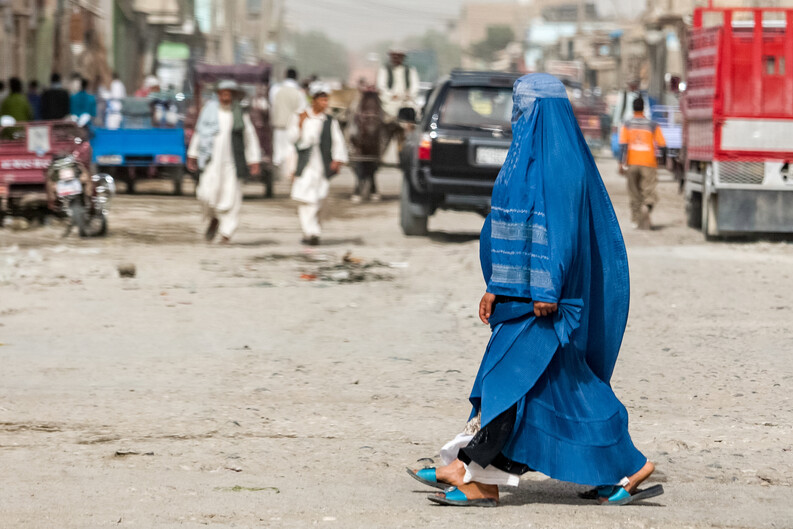 Two women wearing burqa cross a busy unpaved street in Kabul, Afghanistan, with vehicles and other pedestrians visible in the background