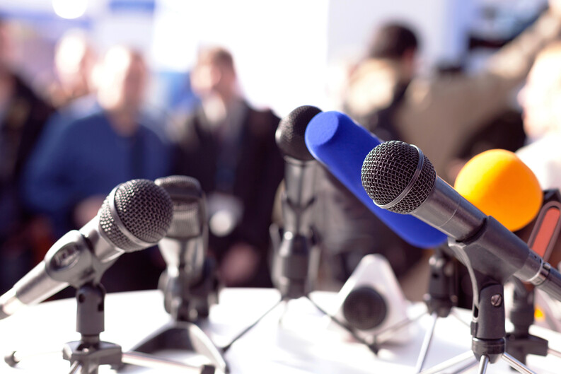 A group of microphones on a table with a crowd in the background.
