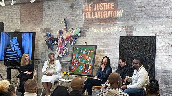 Justice Collaboratory panelists sit talking with artwork behind them and a brick wall with Justice Collaboratory lettering projected onto the wall