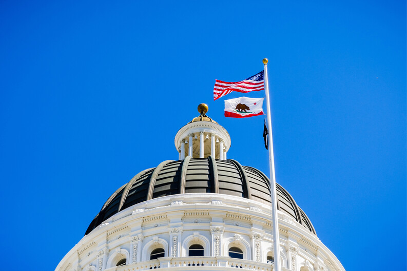 The dome of the California State Capitol against a blue sky with the U.S. and state flags waving in the breeze