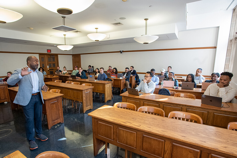 A professor lectures to a classroom with students seated at wooden tables