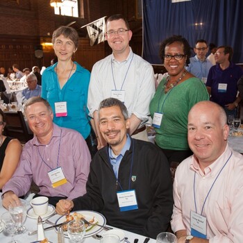 A group of alumni seated at a dinner table at a reception with more alumni standing behind them