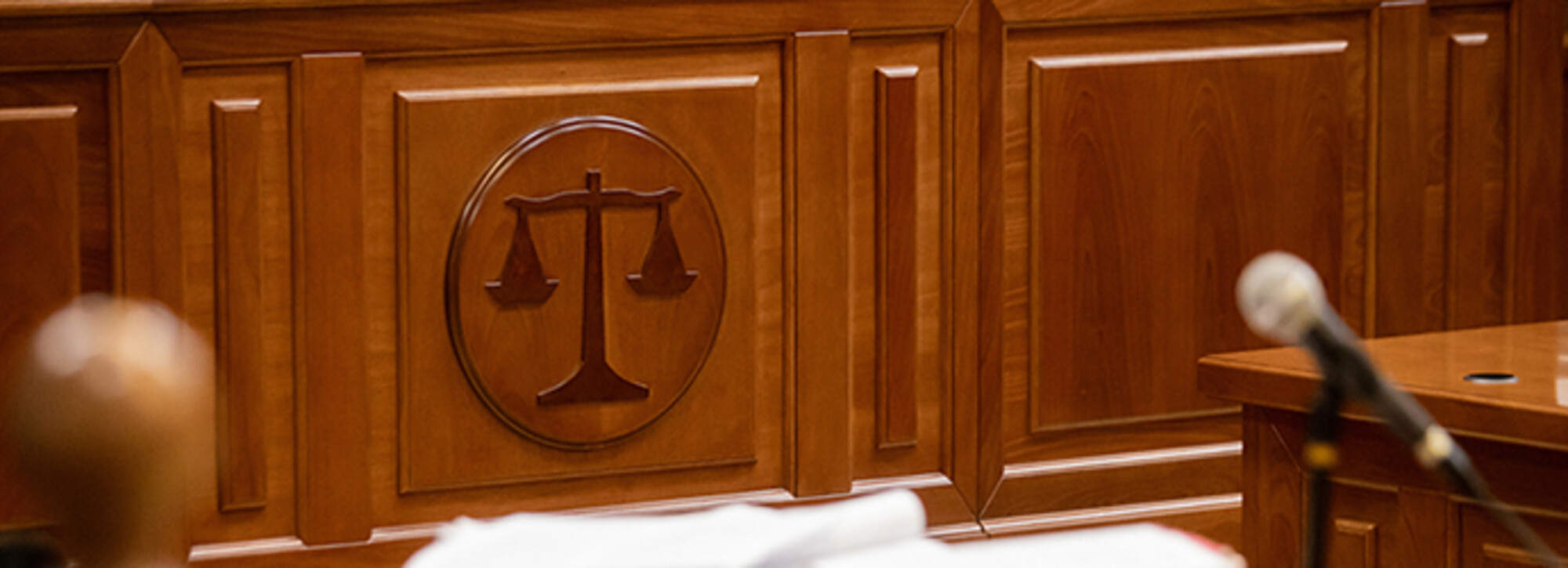 The front of a judge's bench with scales of justice depicted in the wood paneling. A table and microphone are in the foreground 