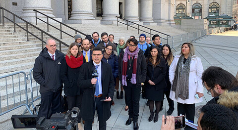A group of people standing on courthouse steps; a man in front holds a microphone and speaks to a camera